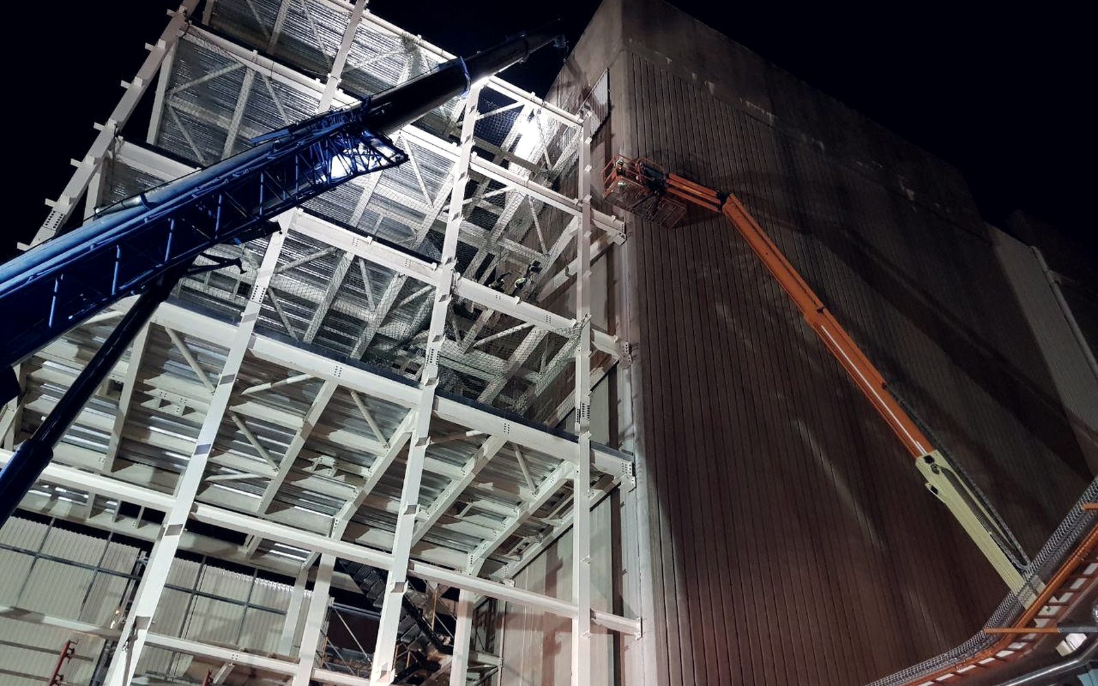 Structural steel frame under erection at night, with a mobile crane and elevated work platform alongside an industrial plant building